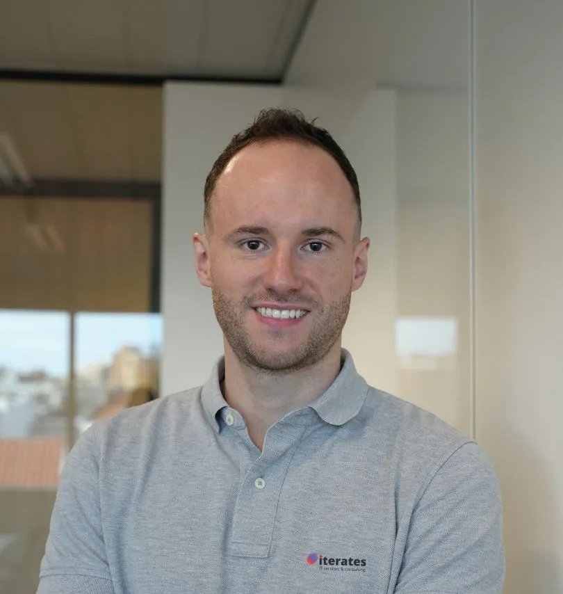 A man with short brown hair and a trimmed beard smiles at the camera. He is wearing a light grey polo shirt with an "iterates" logo. The background shows an office of the Brussels Web Agency with large windows and blurred outside light.